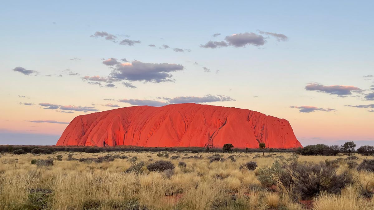 a large red rock in the middle of a dry field with Uluru in the background